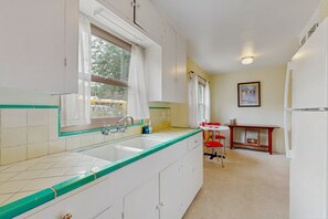 Eine Küche mit Spüle, Schränken, einem Fenster mit Vorhängen, einem Tisch mit Stühlen und einem Kühlschrank.