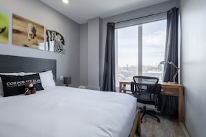 Una habitación de hotel con cama, un escritorio y una silla, una ventana con cortinas y una obra de arte en la pared.