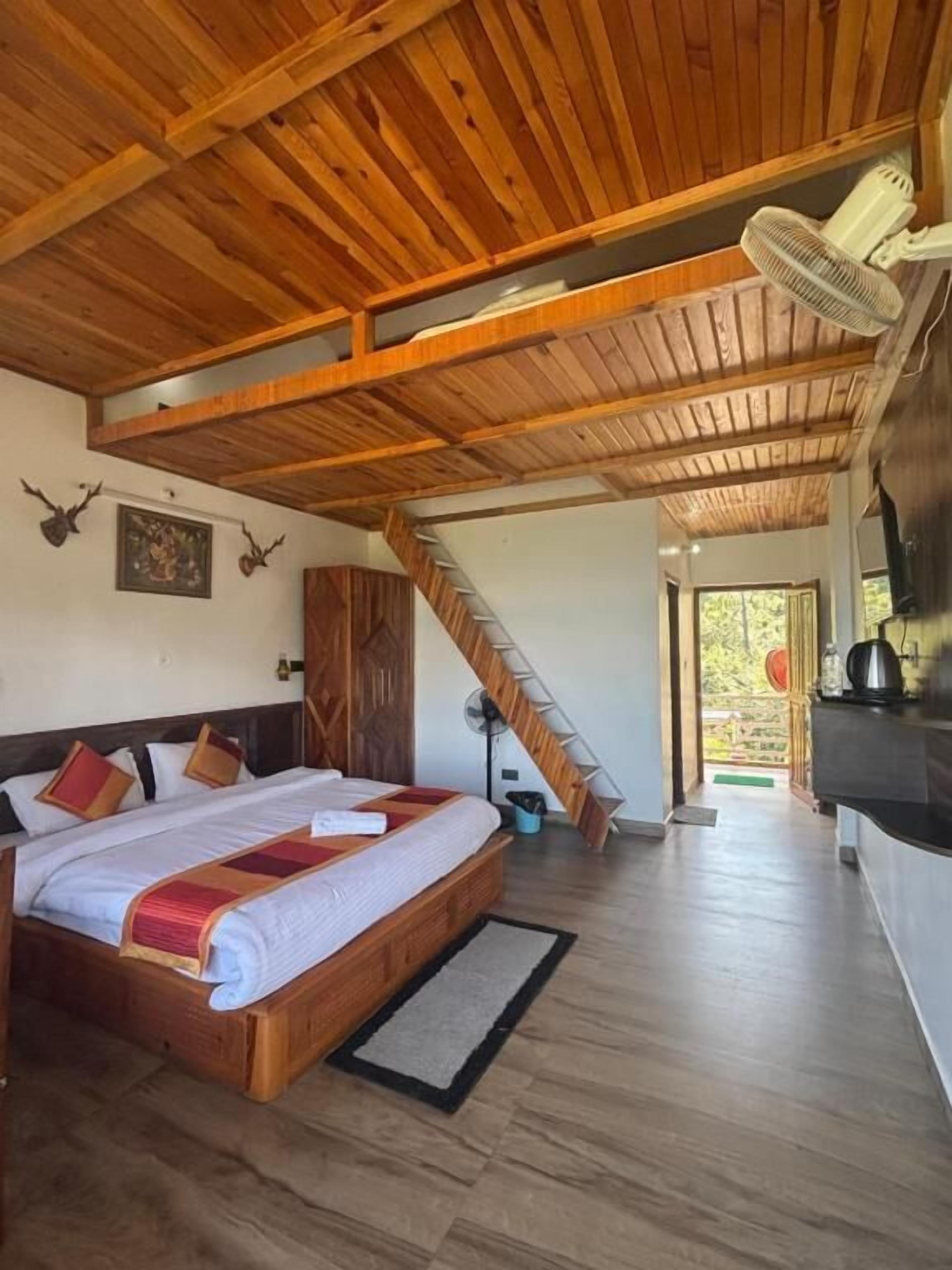 Una habitación con techo de madera, una cama con edredón blanco, cabecera de madera, un ventilador, una escalera y vistas al exterior.