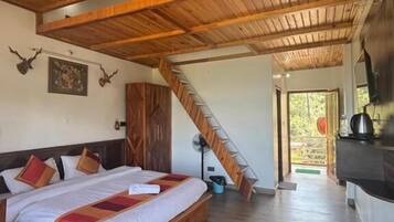 Una habitación con techo de madera, una cama con edredón blanco, cabecera de madera, un ventilador, una escalera y vistas al exterior.