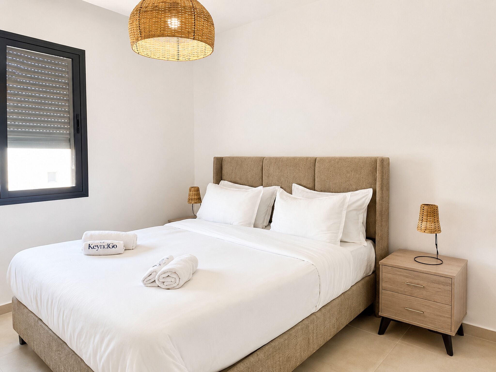 A neatly made bed with white linens and pillows, a bedside table with a lamp, and a window with blinds.