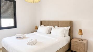 A neatly made bed with white linens and pillows, a bedside table with a lamp, and a window with blinds.