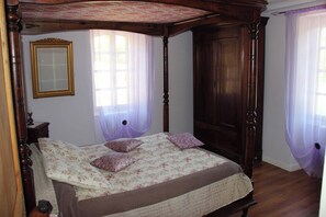 Uma cama de dossel com colcha e almofadas florais, um guarda-roupa de madeira, um espelho na parede e cortinas de tecido leve.