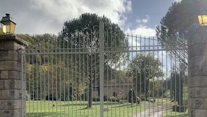 Een toegangspoort met een stenen muur, een metalen hek en een pad dat naar een gebouw leidt.