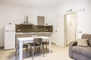 A compact kitchen with a white refrigerator, a marble countertop, and a modern stove. There is a dining table with chairs and a sofa in the adjacent living area. - Nova Suite - Apartment 100 (Calatafimi)