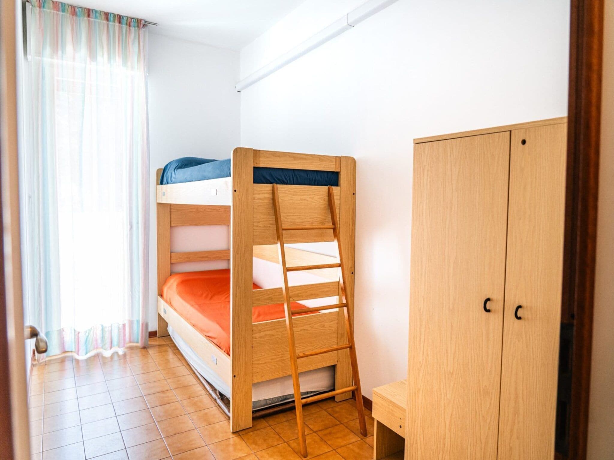 Ein Etagenbett mit einer Stehle, ein Holzschrank und ein kleiner Tisch in einem Zimmer mit Fliesenboden und einem Fenster mit leichtem Vorhang.