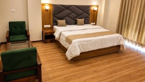 A hotel room with a large bed, a green armchair, a glass coffee table, wooden flooring, and a headboard with wall-mounted lights. - Omya Mountain Retreat Manali (Manali)