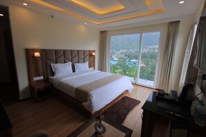 A hotel room with a large bed, a desk with a computer, and a view of a cityscape through the window. - Omya Mountain Retreat Manali (Manali)