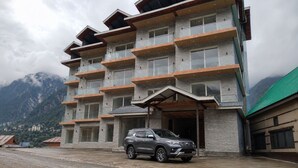 A multi-story building with balconies, a car parked in front, and mountains in the background. - Omya Mountain Retreat Manali (Manali)