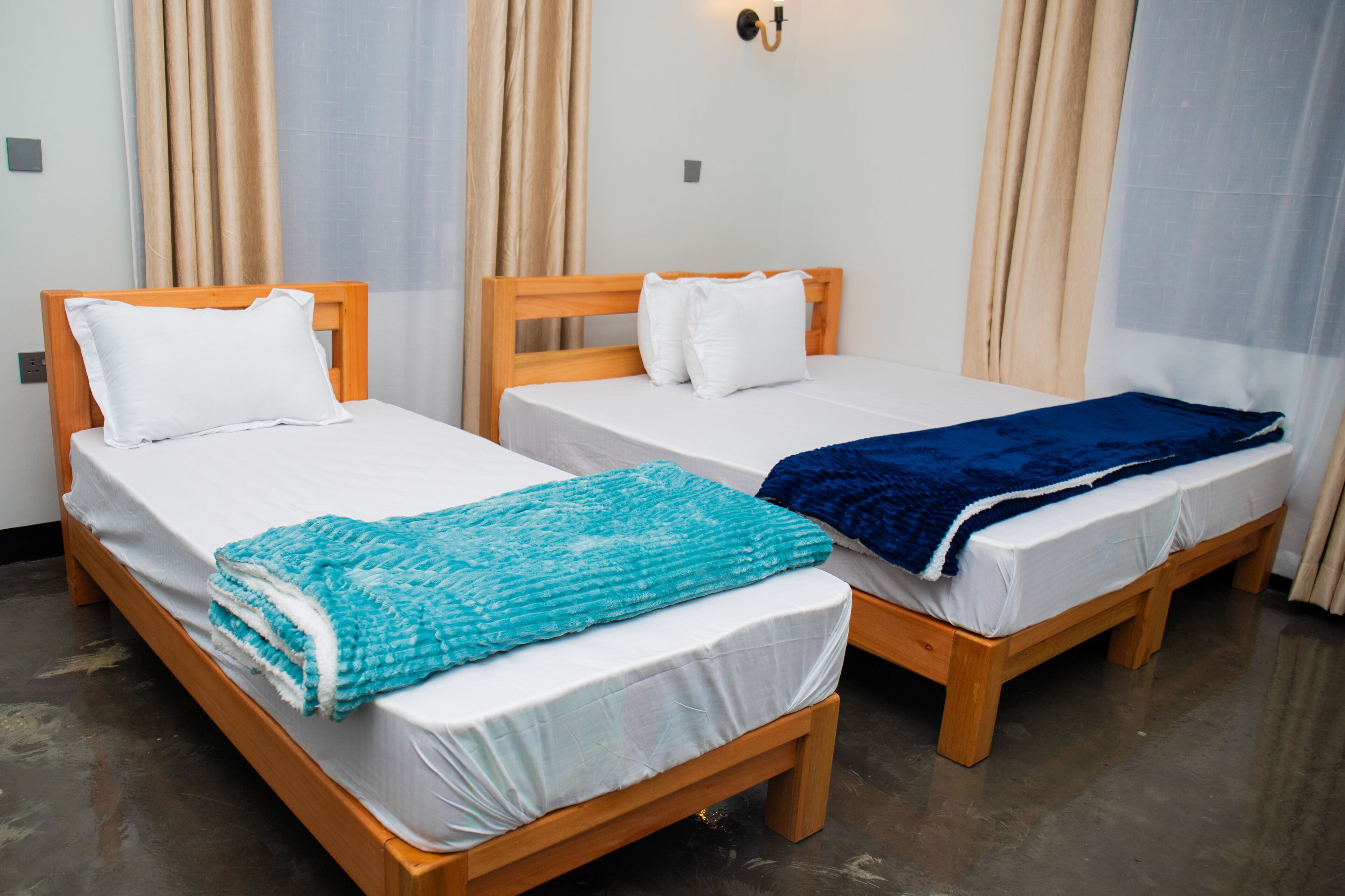 Two single beds with wooden frames and white bedding, a blue blanket on one bed, and beige curtains on the windows.