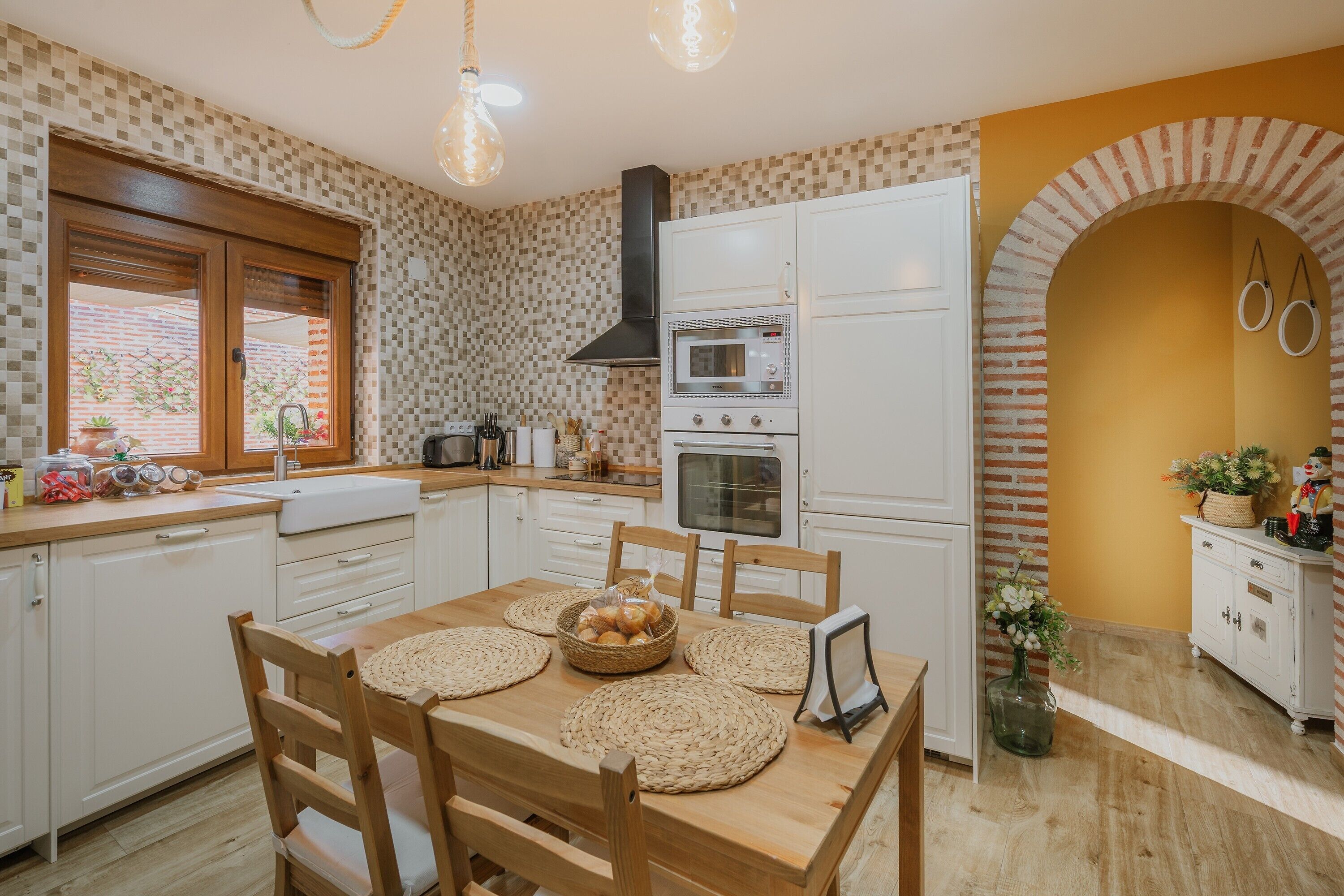 Una cocina con armarios blancos, una mesa de comedor de madera con individuales, una cesta de pan y una lámpara colgante.