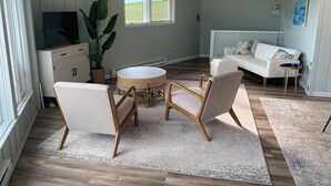 Ett rum med ett runt träbord, två stolar, en vit soffa, en tv, en stor växt och ett fönster med utsikt över grönområden.