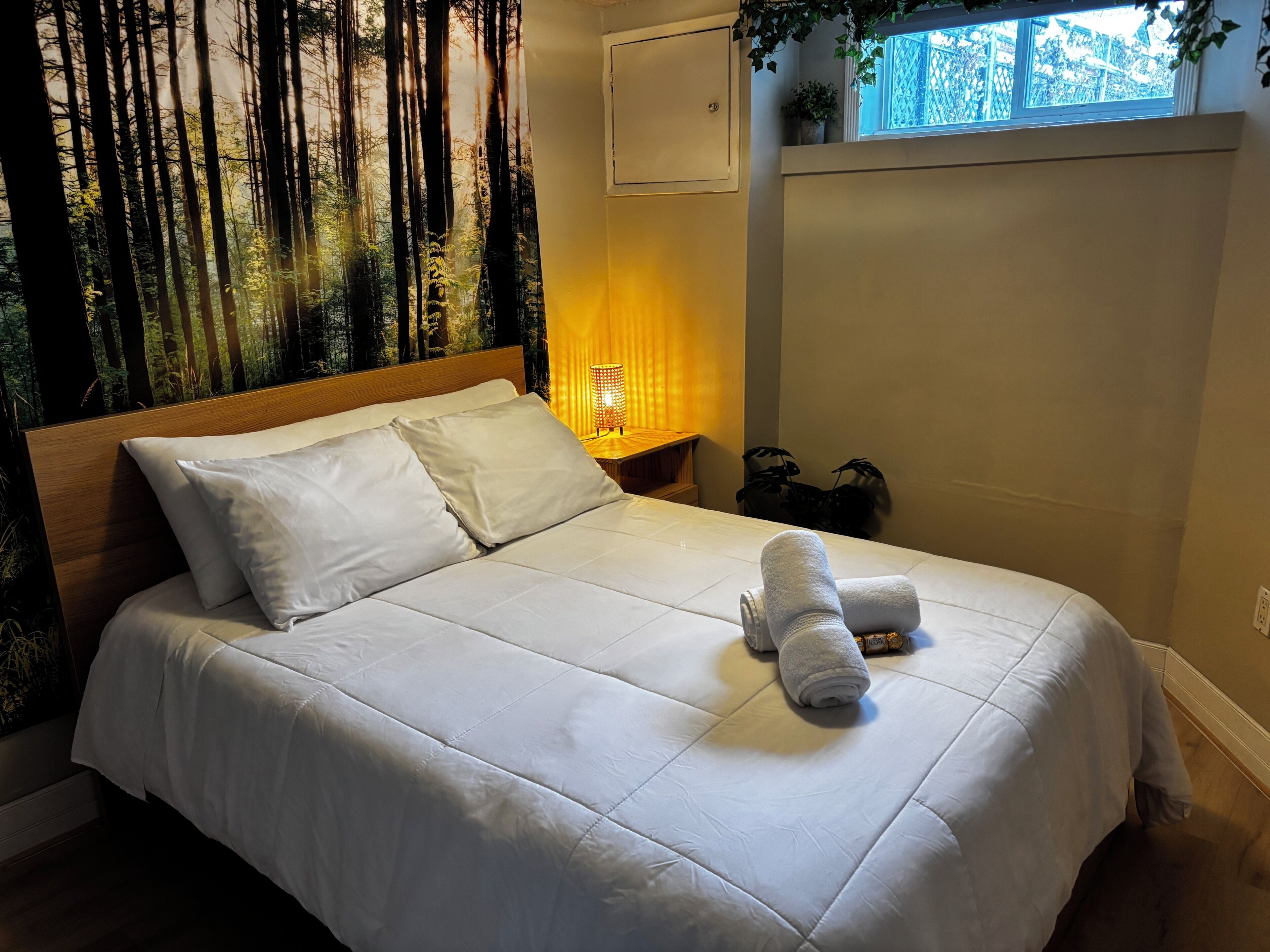 Een netjes opgemaakt bed met witte lakens en twee opgerolde handdoeken, een bedlamp en een raam met uitzicht op bomen.