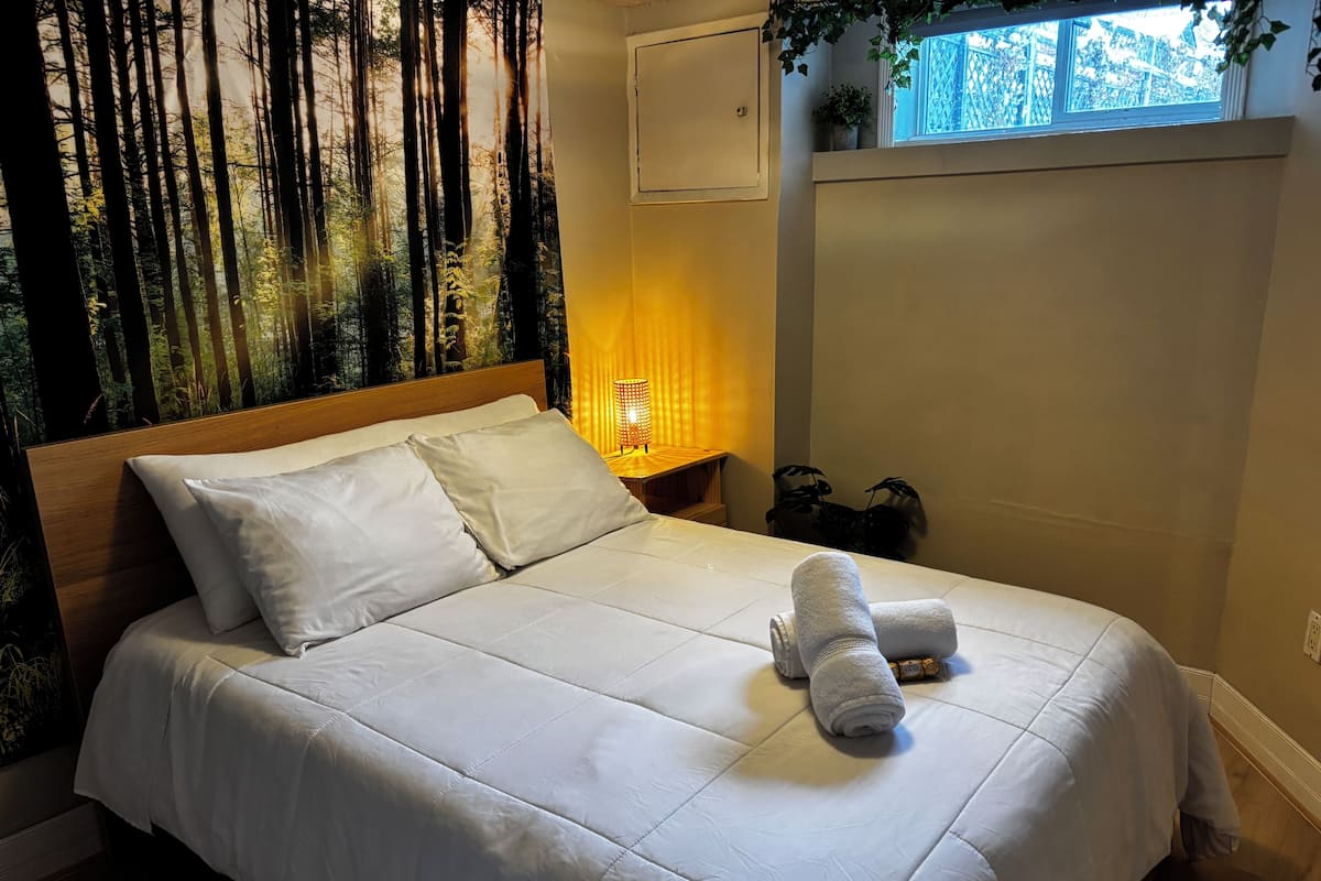 Una cama bien tendida con sábanas blancas y dos toallas enrolladas, una lámpara de noche y una ventana con vistas a árboles.