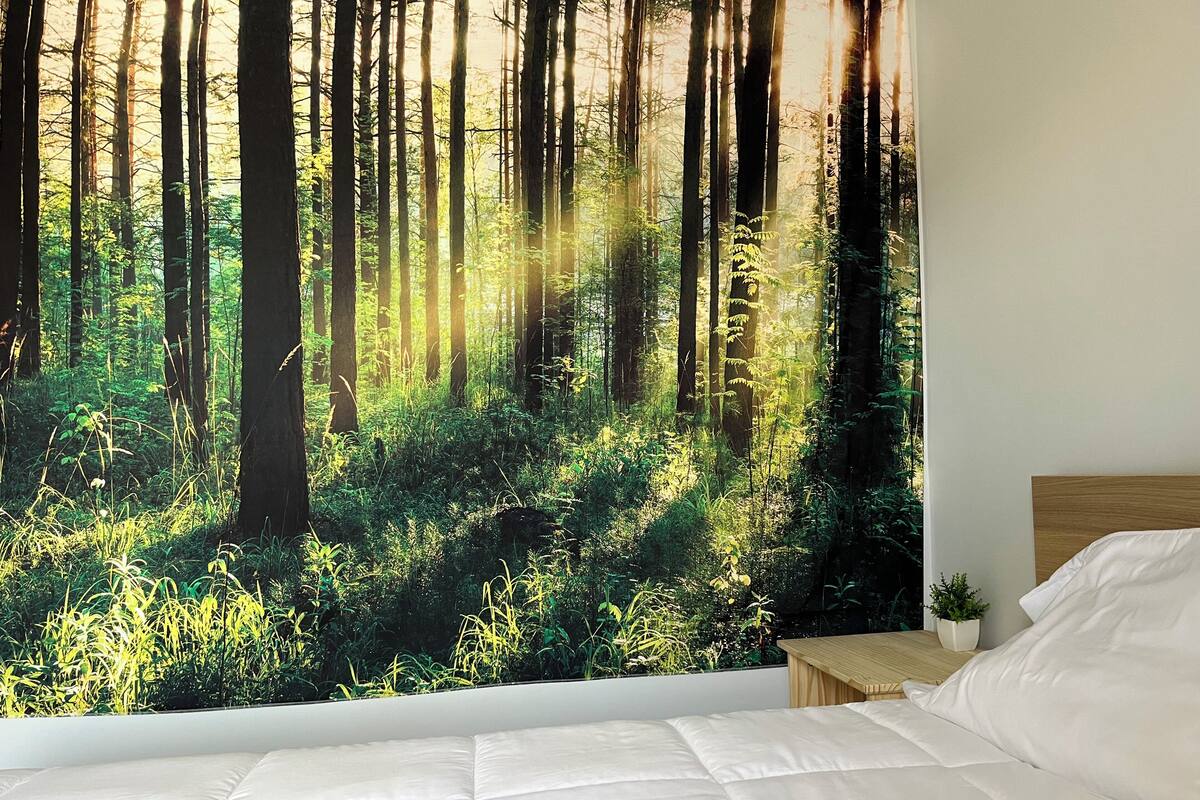Un dormitorio con un mural grande de temática forestal, una cama bien hecha y una mesita de noche pequeña con una planta.
