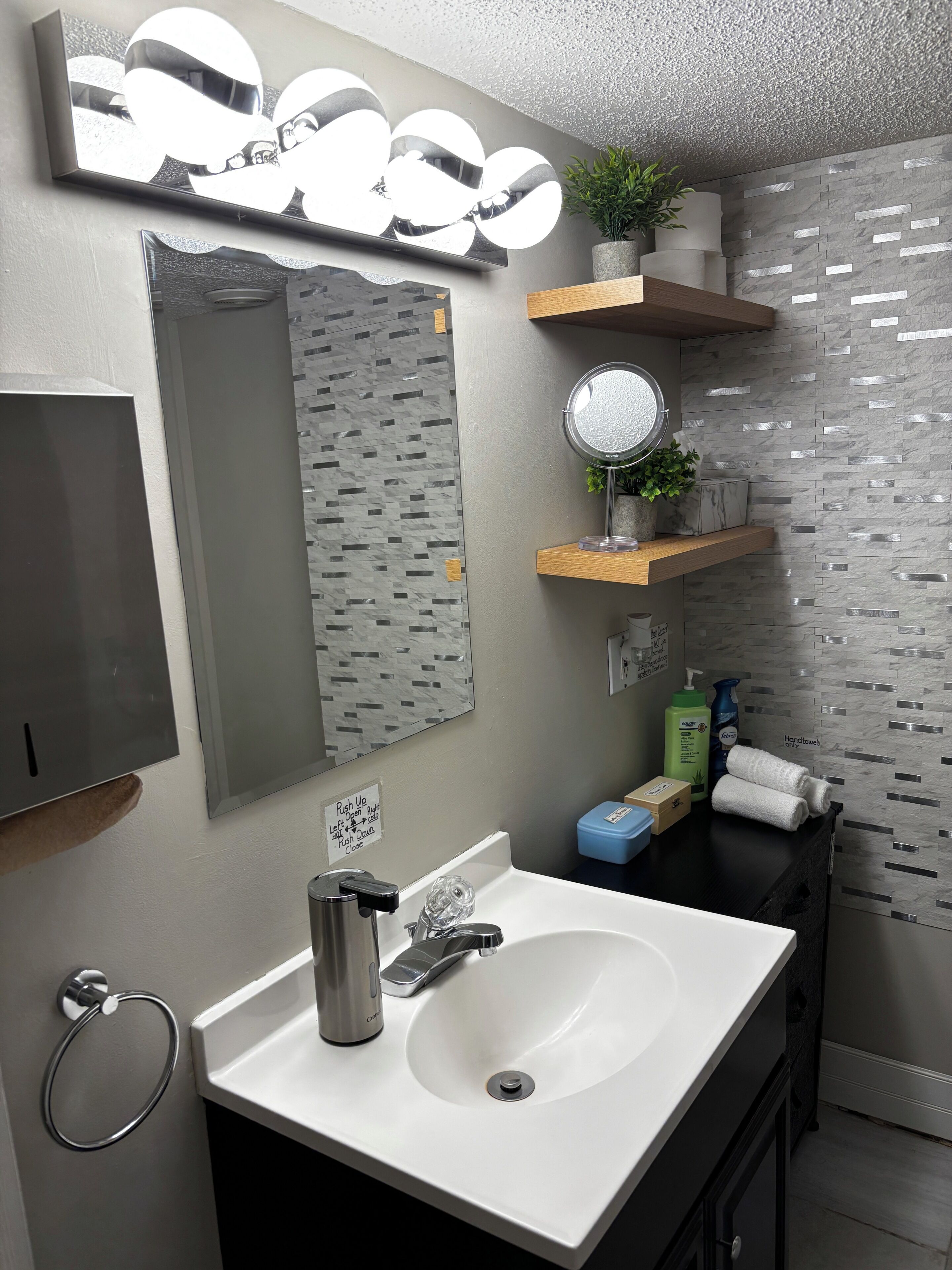 Un baño moderno con lavamanos blanco, grifería de acero inoxidable, un espejo con luces y una pared con textura.