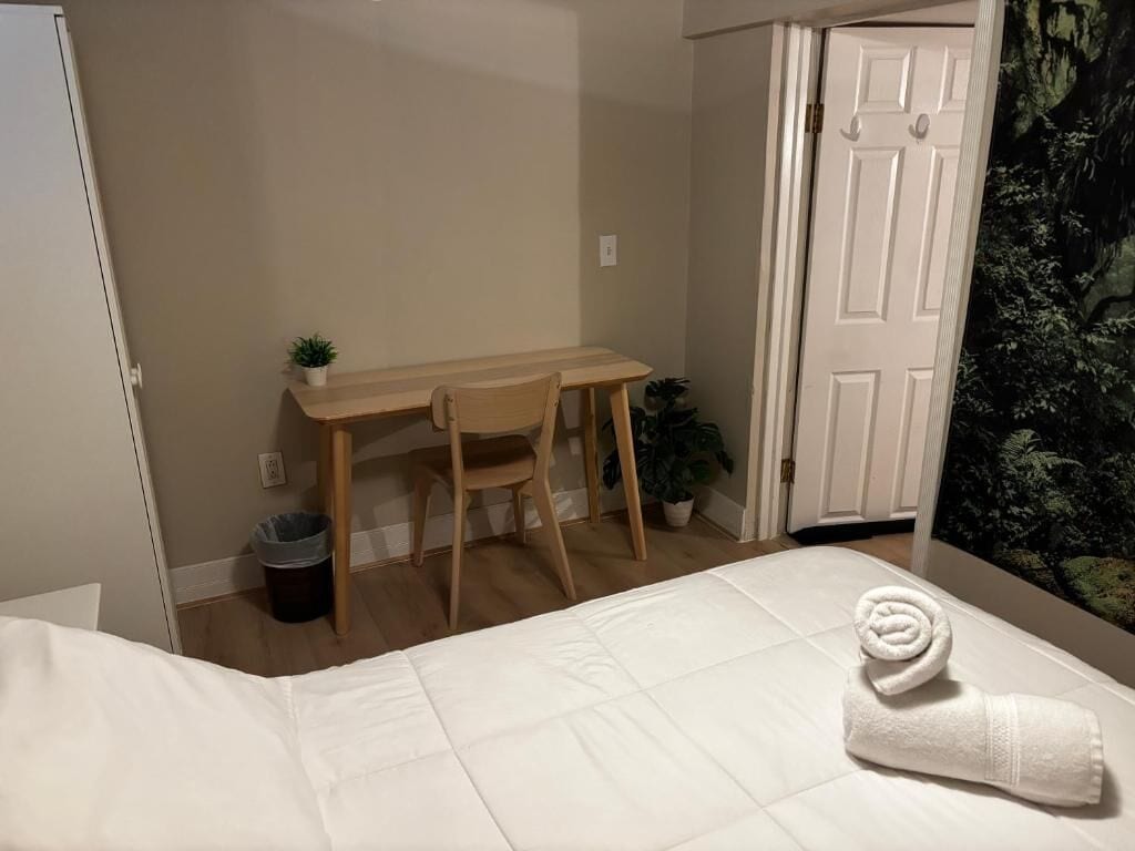A bedroom with a bed, a desk, a chair, a small plant, and a door with a forest mural.