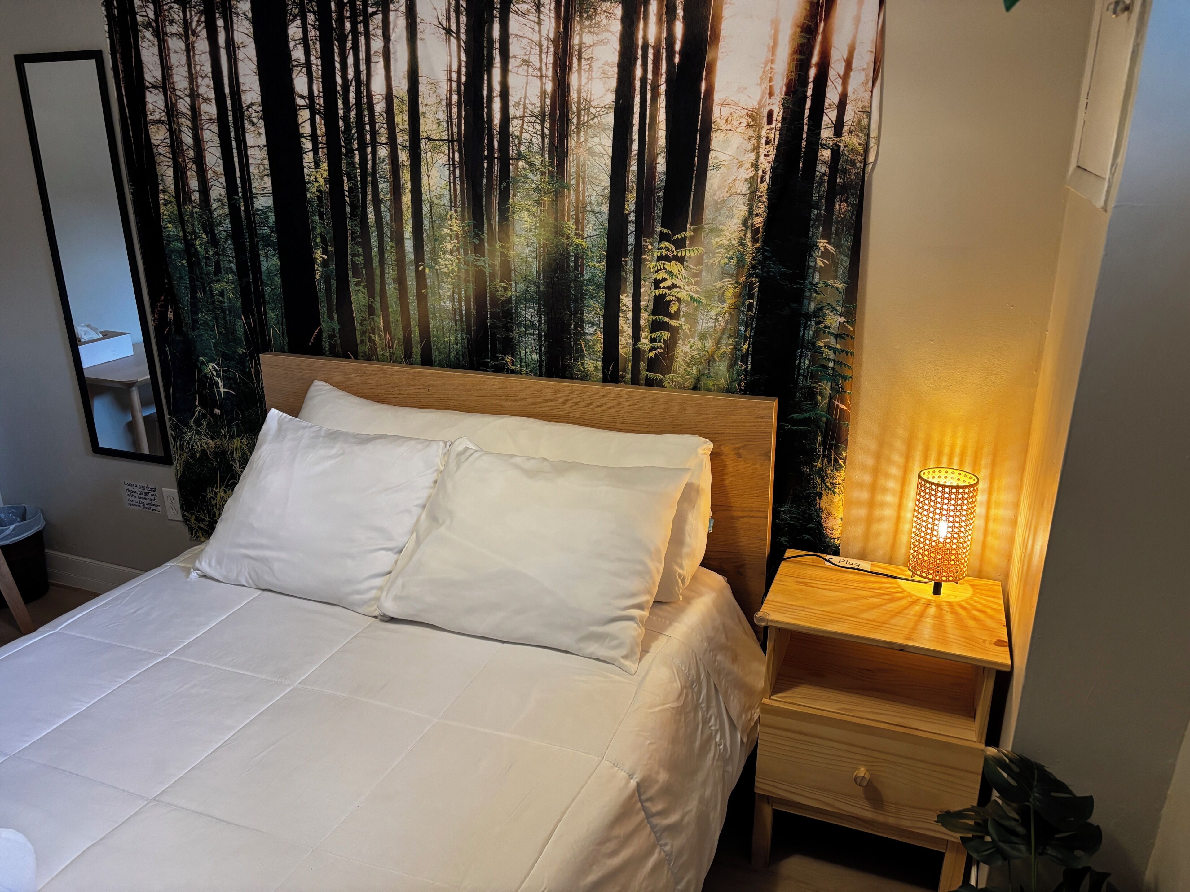 Una cama con ropa de cama blanca, cabecera de madera, mesita de noche con lámpara y un tapiz mural con motivos forestales.
