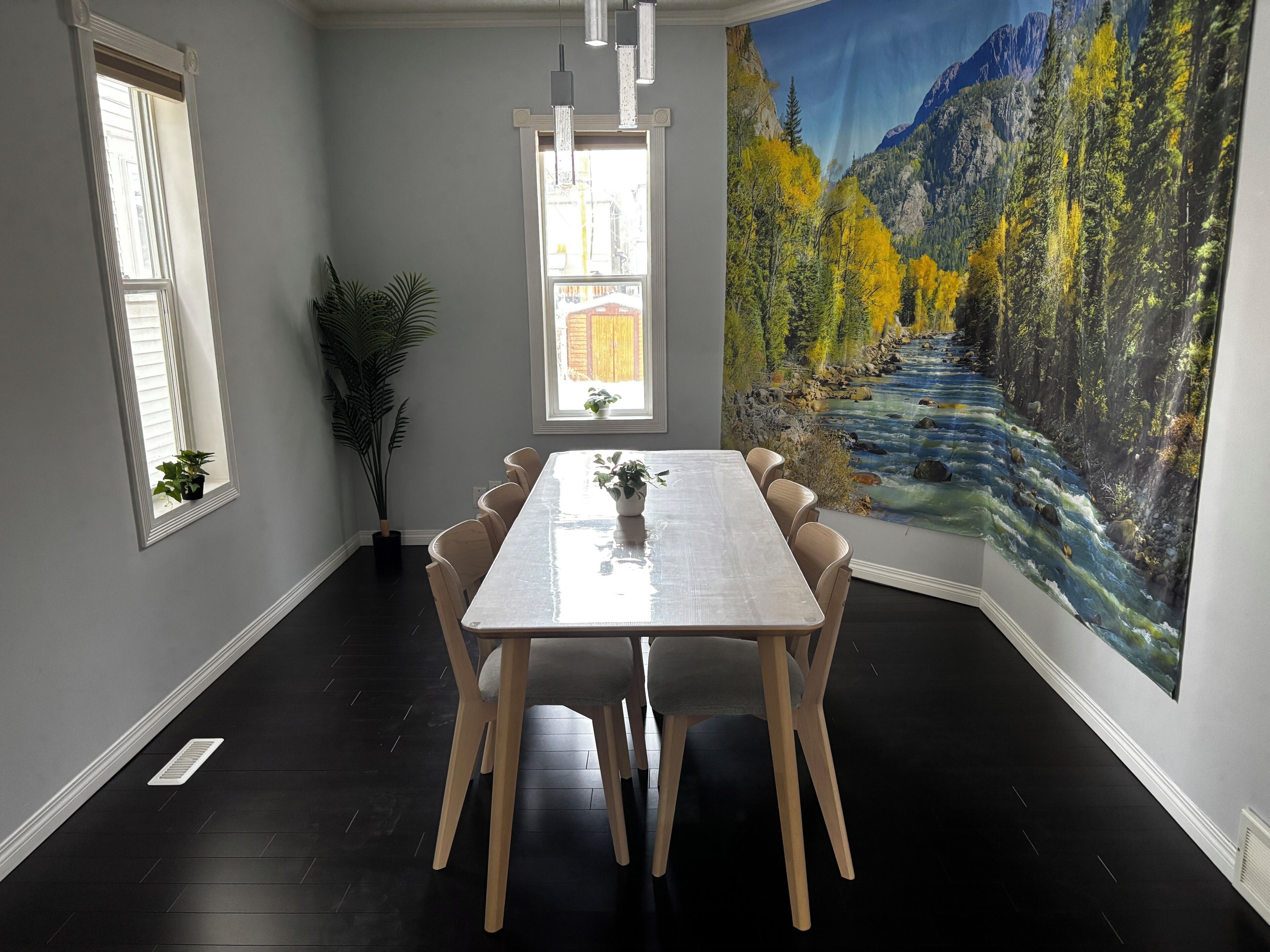 Un comedor con una mesa larga, sillas, una planta en maceta y un gran mural de un bosque y un río.