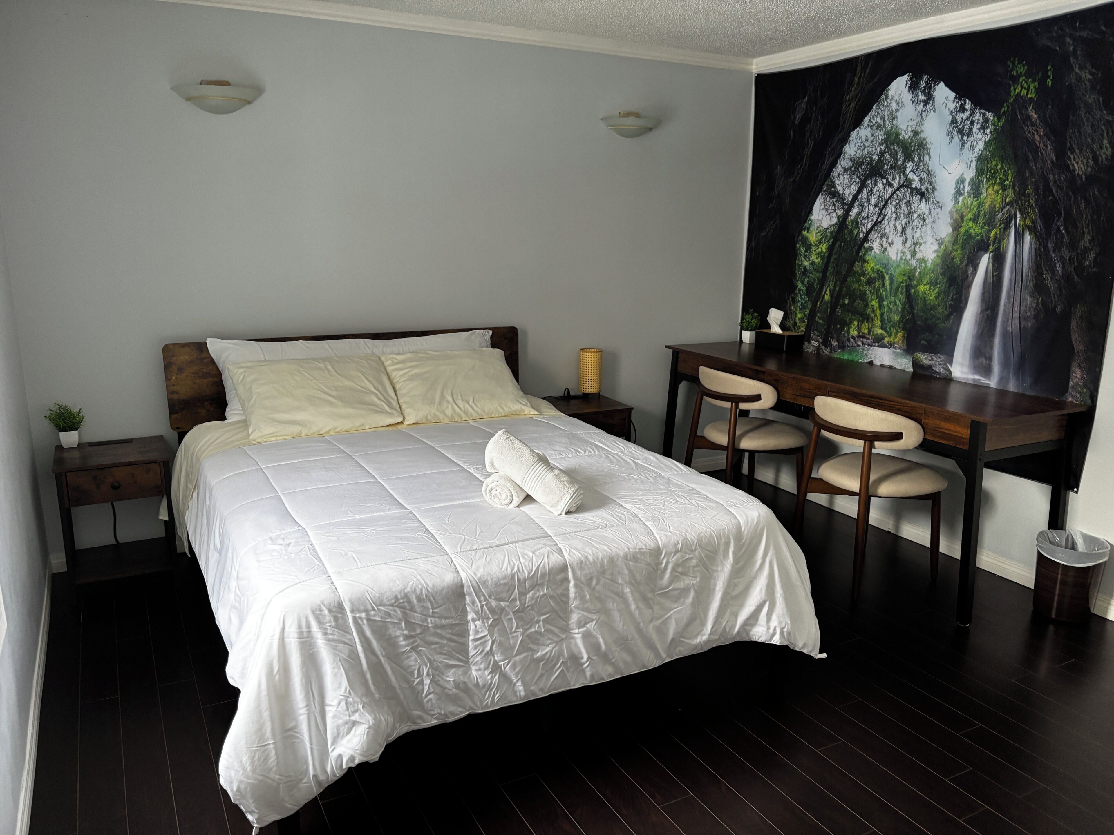Une chambre à coucher comprenant un lit, un bureau, deux chaises, une petite table avec une lampe et un luminaire fixé au mur.
