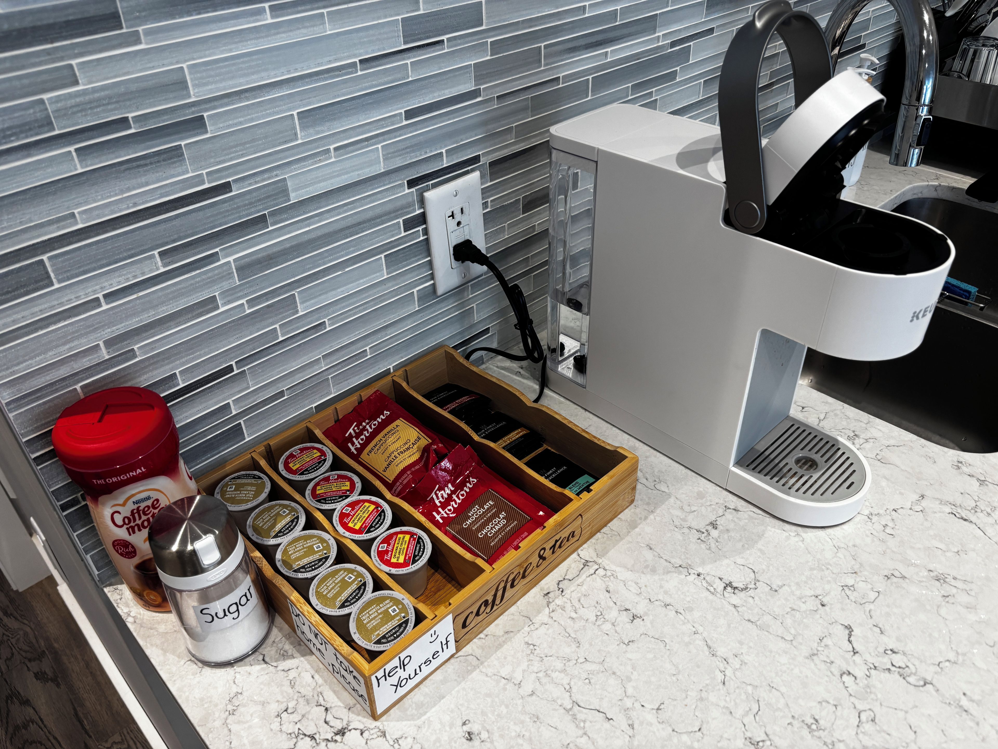 Una encimera de cocina con una cafetera, un tazón de azúcar y una caja de cápsulas de café.