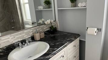 A bathroom with a white sink, marble countertop, and a mirror above it. There are shelves with towels and a potted plant.