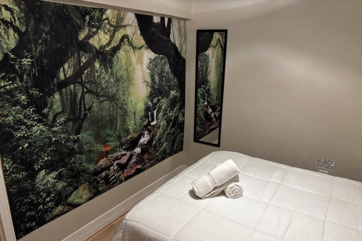 Un dormitorio con un amplio mural de un bosque en la pared, una cama bien hecha con una toalla enrollada, y un pequeño cuadro enmarcado en la pared.
