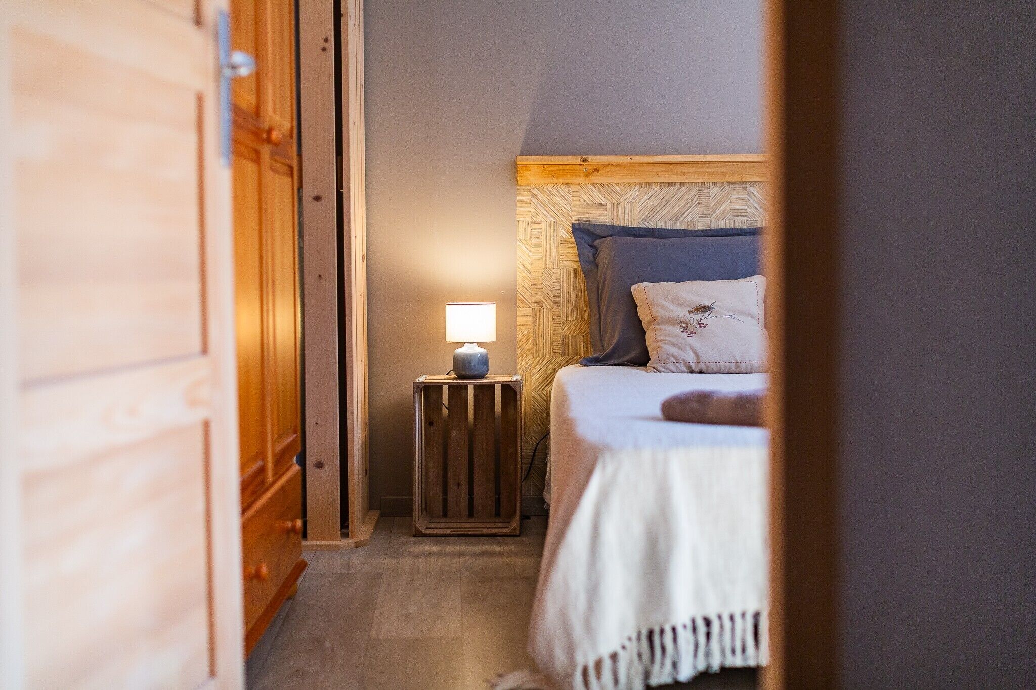 Un couloir étroit mène à une chambre à coucher qui contient un lit, une table de chevet avec une lampe et une armoire en bois.