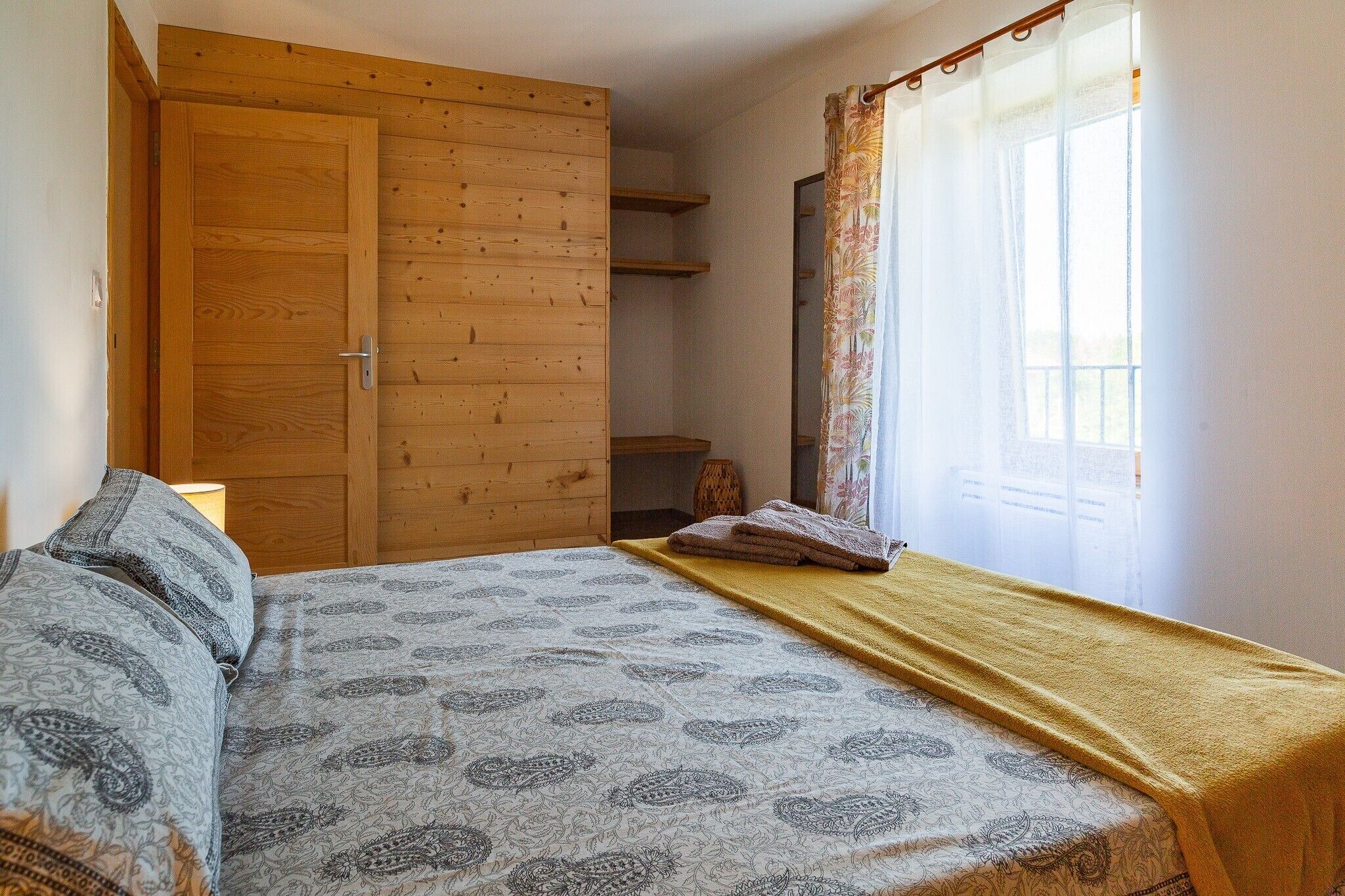 A bedroom with a bed, a wooden wardrobe, a window with curtains, and a bedside lamp.