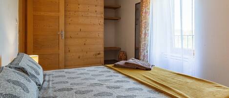 A bedroom with a bed, a wooden wardrobe, a window with curtains, and a bedside lamp.