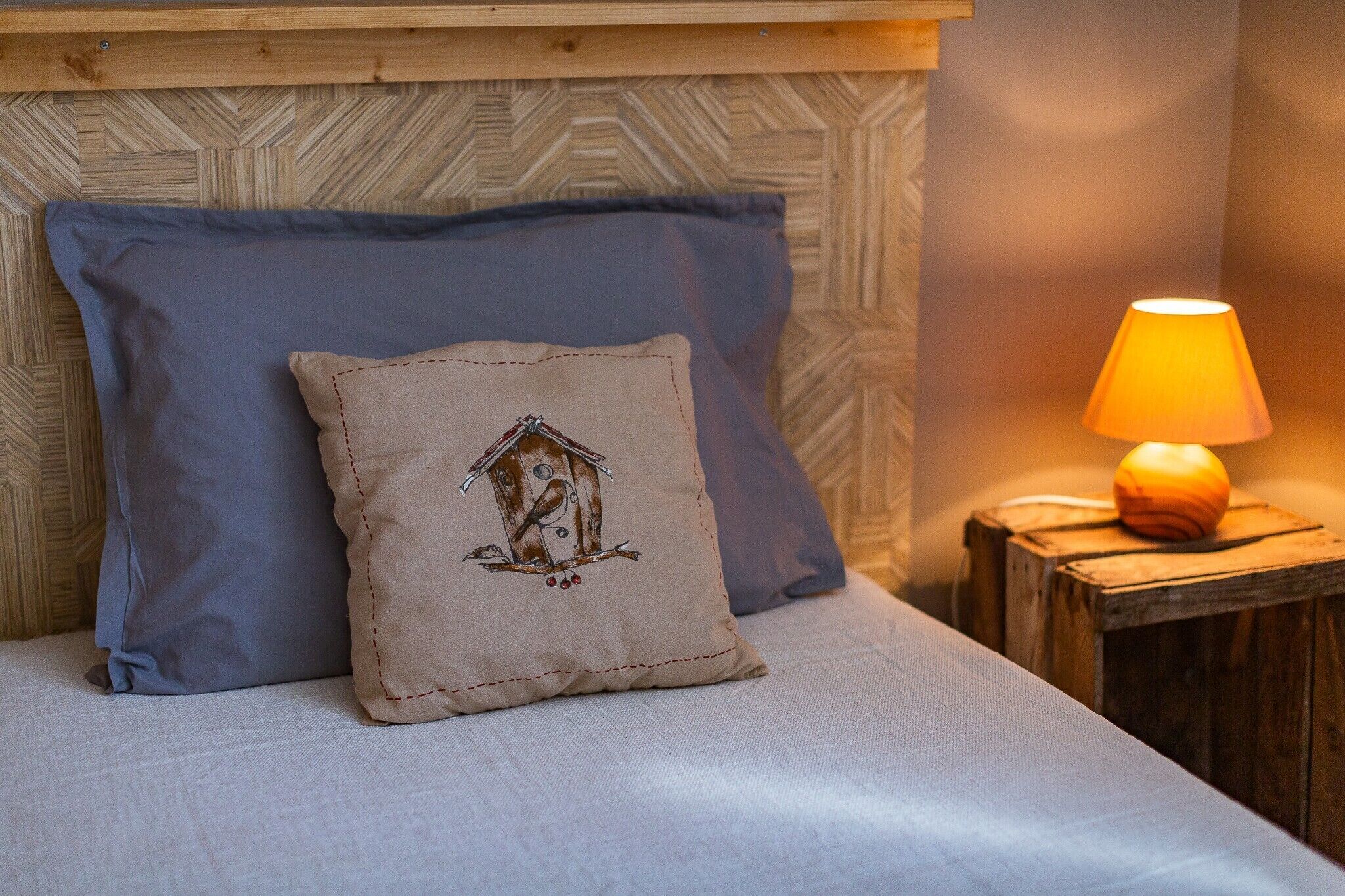 Ein Bett mit blauem und beigem Kissen, wobei eines mit Häuserstickerei verziert ist, ein Holz-Kopfteil und ein kleiner Tisch mit einer Lampe.