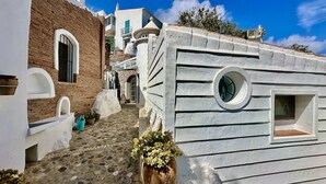 Uma rua de paralelepípedos estreita, ladeada por edifícios com arquitetura tradicional, incluindo uma parede de pedra e um edifício com janela circular.