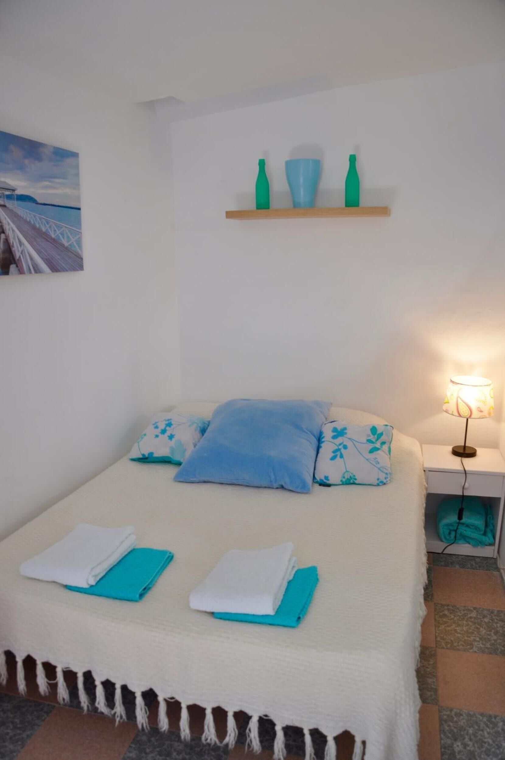 Un lit avec une couverture blanche et bleue, un oreiller bleu et un oreiller blanc. Il y a une petite étagère au-dessus du lit, sur laquelle sont posées trois bouteilles.