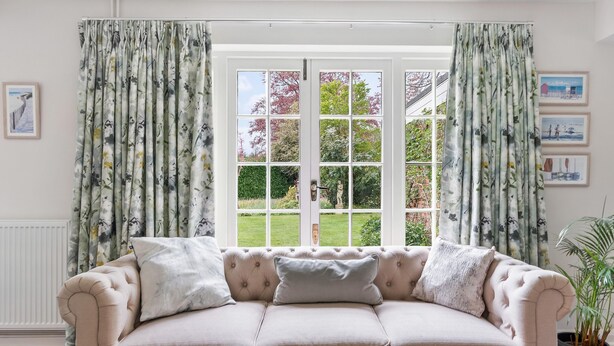 A beige sofa with three cushions, a window with floral curtains, and a view of a garden. - Dairy Close Parking By My Getaways (Uckfield)
