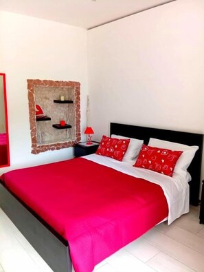 A bed with a red bedspread and pillows, a nightstand with a red lamp, and a wall-mounted shelf with decorative items.
