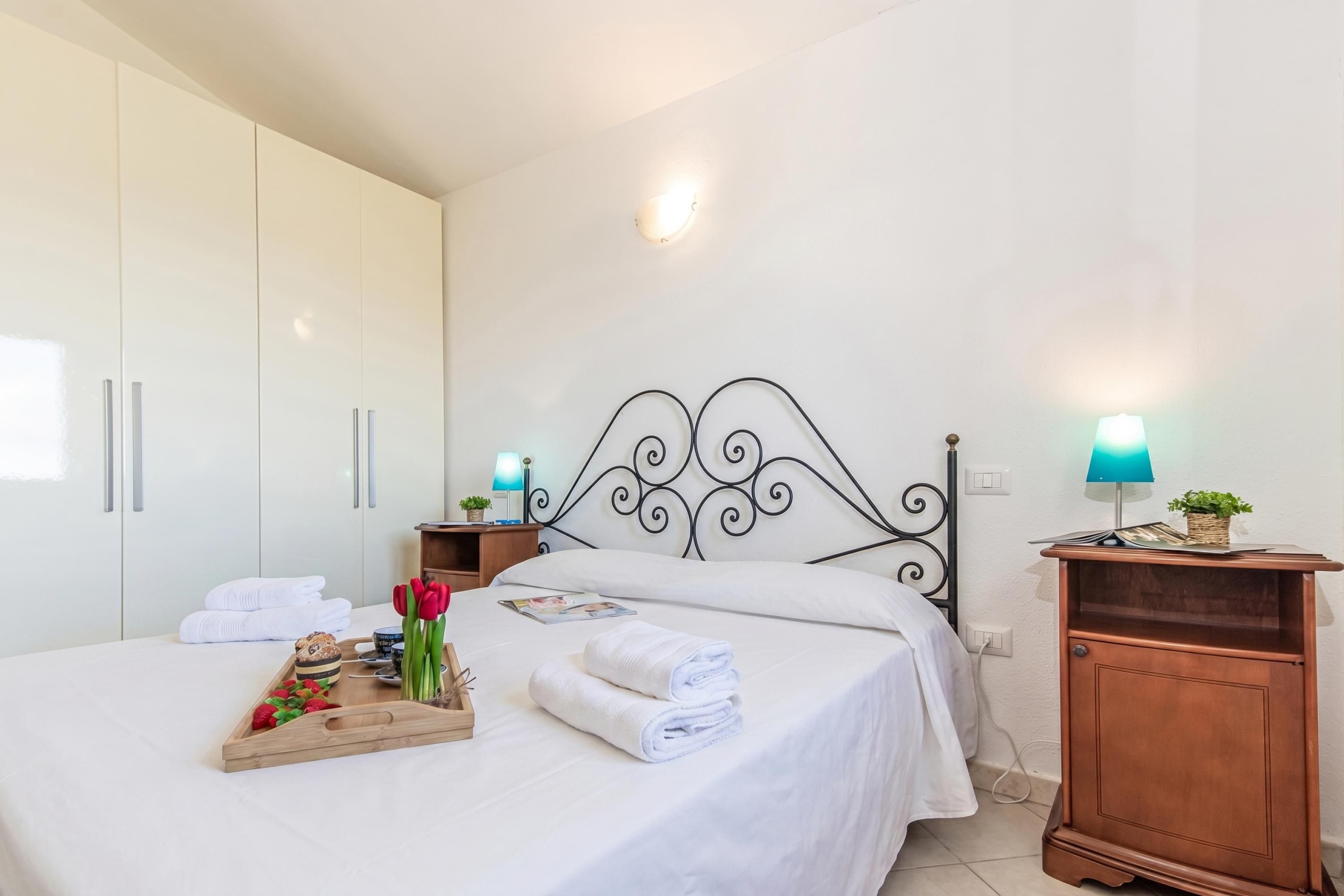 Una cama bien hecha con sábanas blancas, una bandeja con comida y flores, una mesita de noche de madera con una lámpara y un armario con puertas correderas.