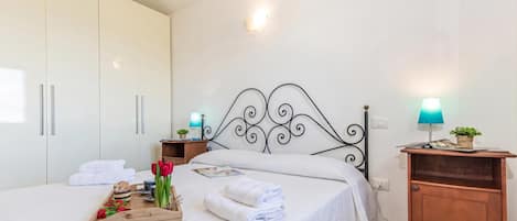 Uma cama bem arrumada com lençóis brancos, um suporte com comida e flores, uma mesa de cabeceira de madeira com abajur, e um guarda-roupa com portas deslizantes.