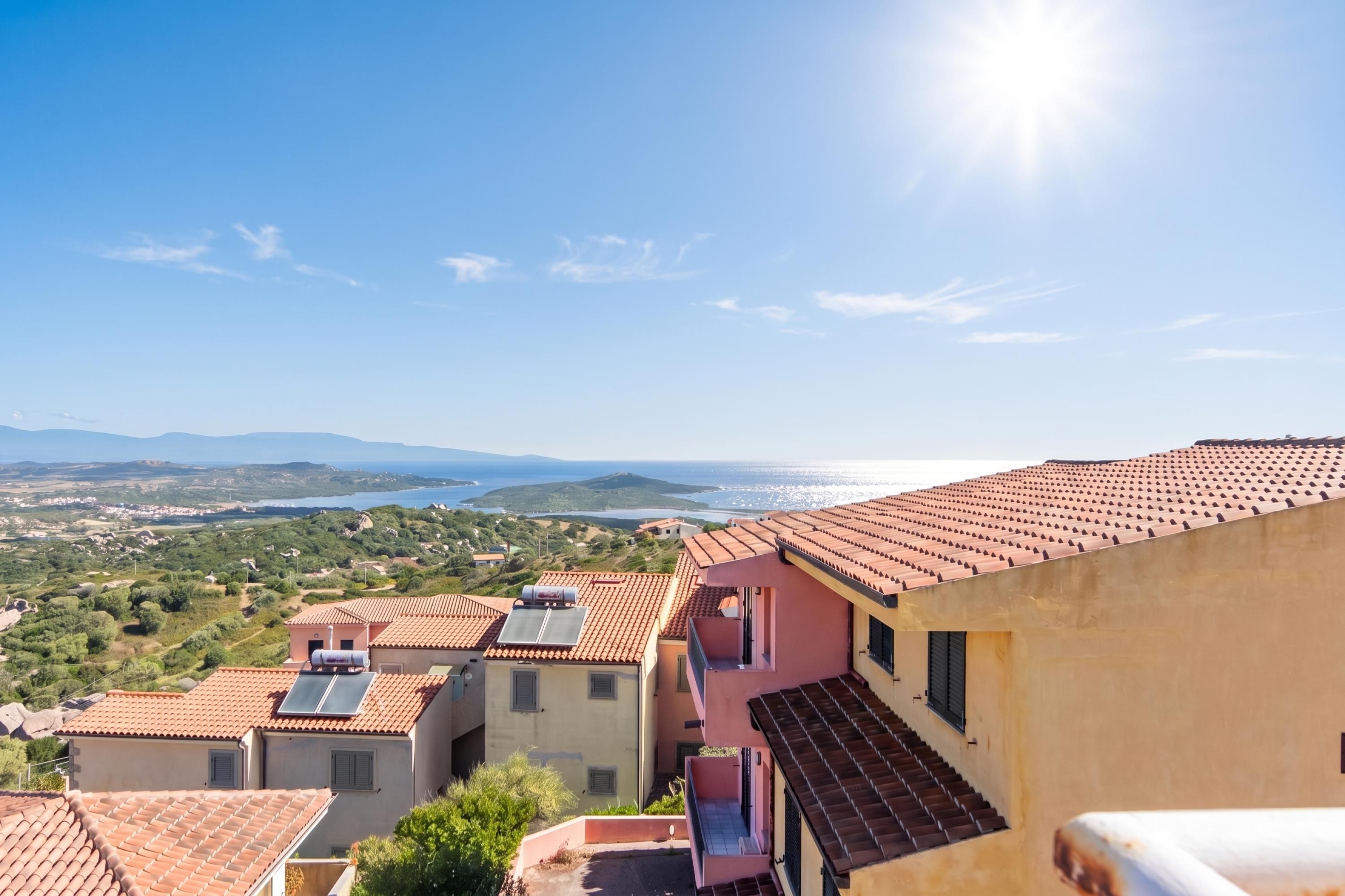 A residential area with terracotta-tiled roofs, a clear sky, and a distant view of a body of water.