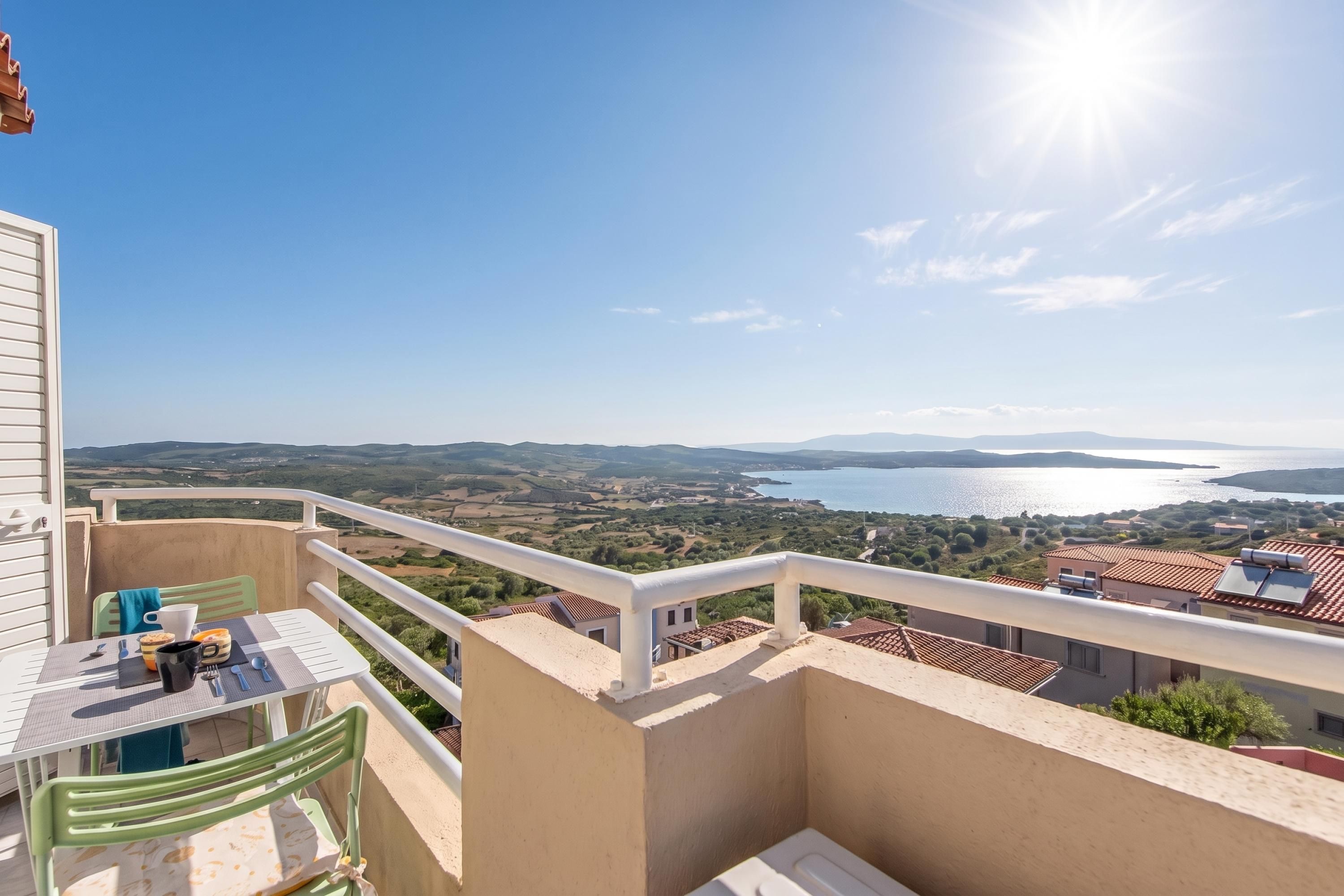 A balcony with a table and chairs, overlooking a coastal landscape with a body of water and distant hills.