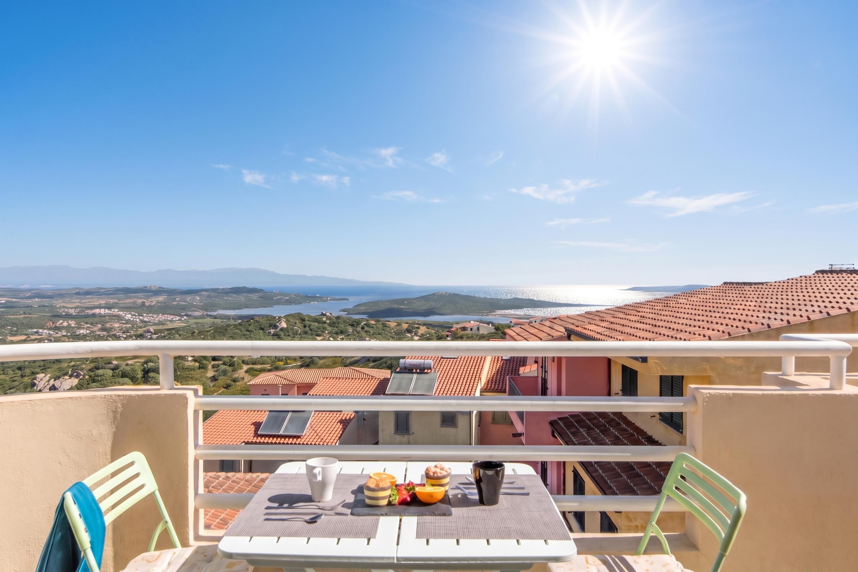 A balcony with a table set for breakfast, overlooking a coastal landscape with terracotta-roofed buildings and a clear sky.