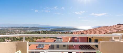 Un balcón con una mesa dispuesta para el desayuno, con vistas a un paisaje costero con edificios de tejados de tejas rojas y un cielo despejado.