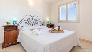 Una cama bien hecha con sábanas blancas, una mesita de noche de madera con una lámpara, una bandeja con comida y flores, y una ventana con vistas al cielo.