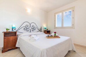 Ein ordentlich gemachtes Bett mit weißen Leinen, ein Nachttisch aus Holz mit einer Lampe, ein Tablett mit Speisen und Blumen sowie ein Fenster mit Blick auf den Himmel.