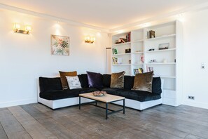 A modern living room with a sectional sofa, a coffee table, a white bookshelf, and wall-mounted lighting. - Le Belvédère - Penthouse, sea & country view (Tourgéville)