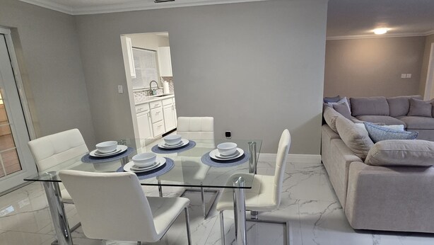 A modern living room with a glass dining table, white chairs, a grey sofa, and a patterned rug.