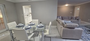 A modern living room with a glass dining table, white chairs, a grey sofa, and a patterned rug.