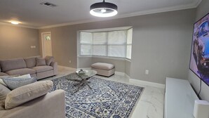 A living room with a beige sofa, a glass coffee table, a patterned rug, a window with blinds, and a TV mounted on the wall.