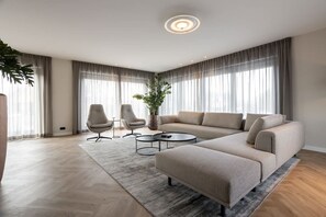 Un salon moderne avec un canapé d’angle, deux fauteuils, une table basse et une grande fenêtre avec des rideaux.