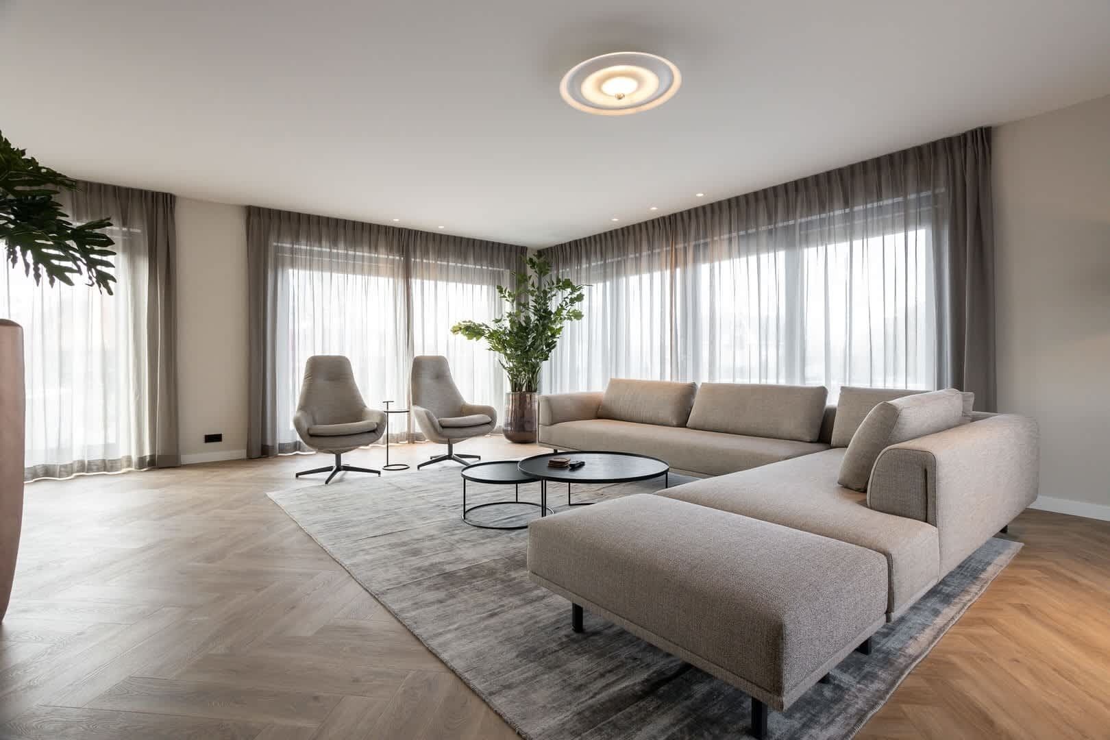Een moderne woonkamer met een hoekbank, twee fauteuils, een salontafel en een groot raam met gordijnen.