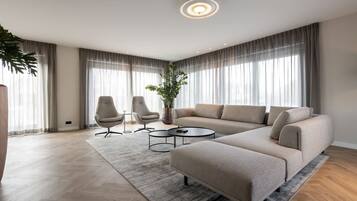 Een moderne woonkamer met een hoekbank, twee fauteuils, een salontafel en een groot raam met gordijnen.